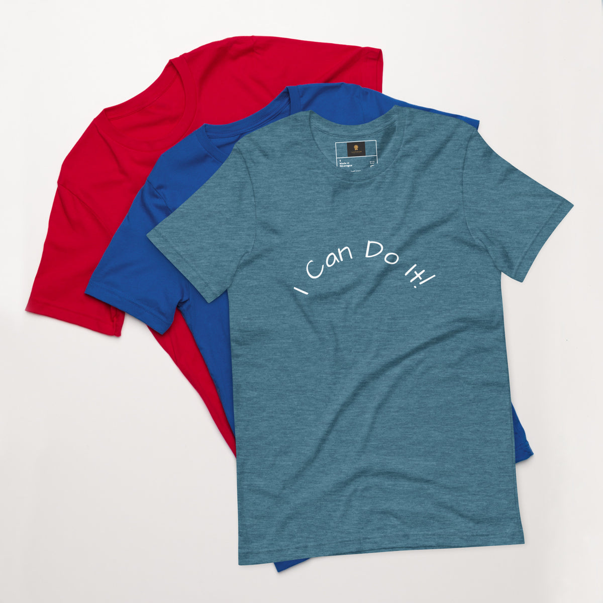 I Can Do It Unisex T-shirt in gray with red and blue shirts stacked in the background.