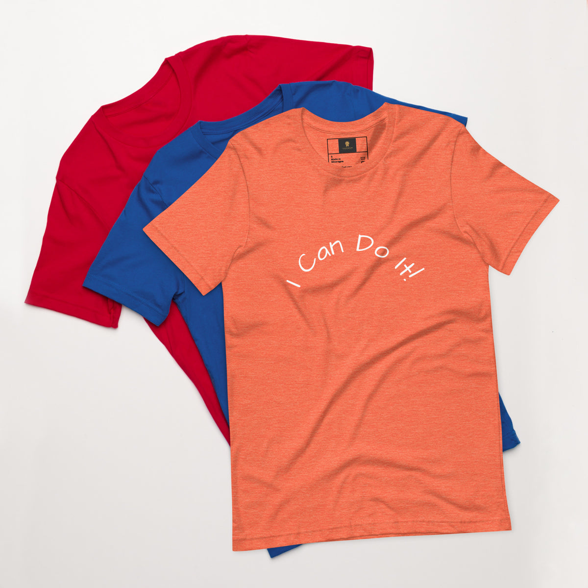 I Can Do It Unisex T-shirt in gray with red and blue shirts stacked in the background.
