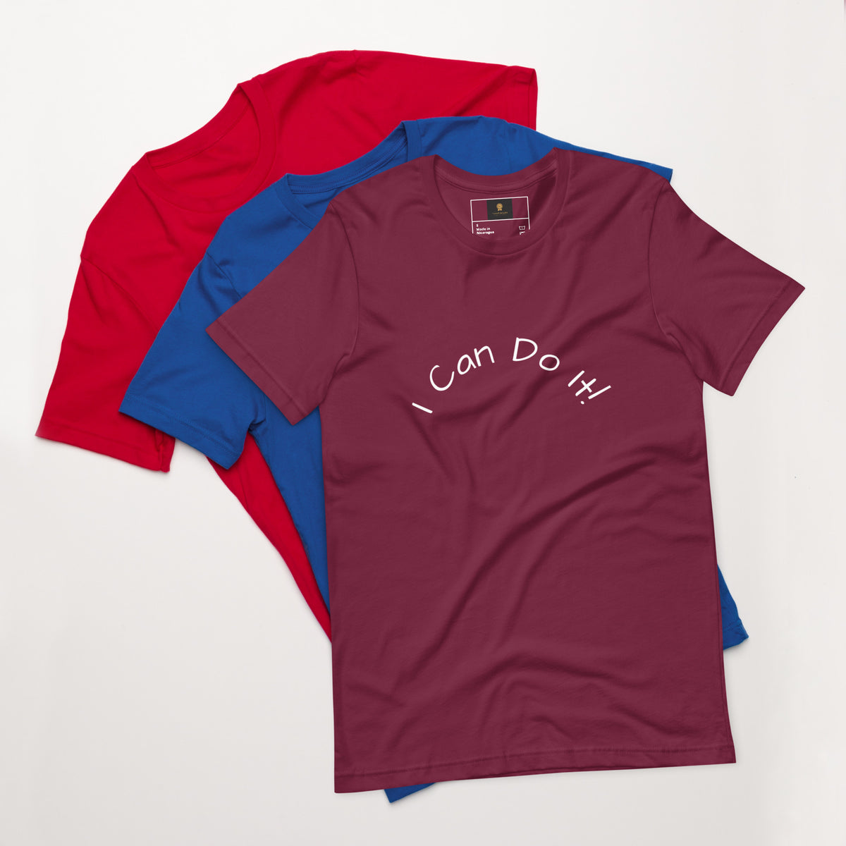 I Can Do It Unisex T-shirt in gray with red and blue shirts stacked in the background.